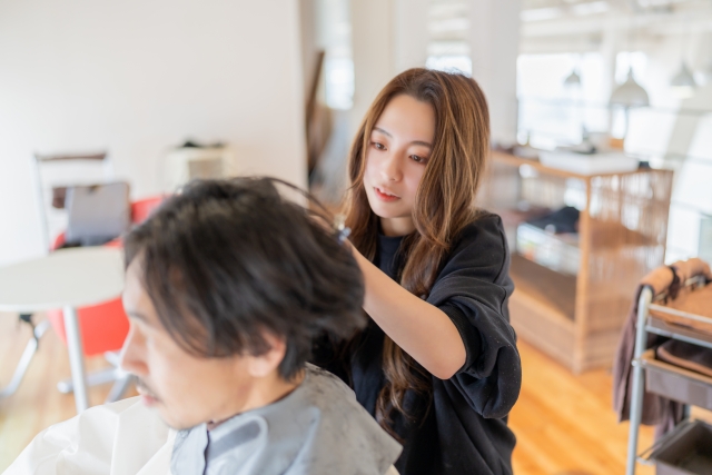 美容師がクライアントの髪を丁寧に整える様子。鋭いまなざしと巧みな手さばきが印象的で、集中した表情には思わず見とれてしまうほど。プロの技術と情熱が伝わる一瞬をとらえた美しいカットシーン。