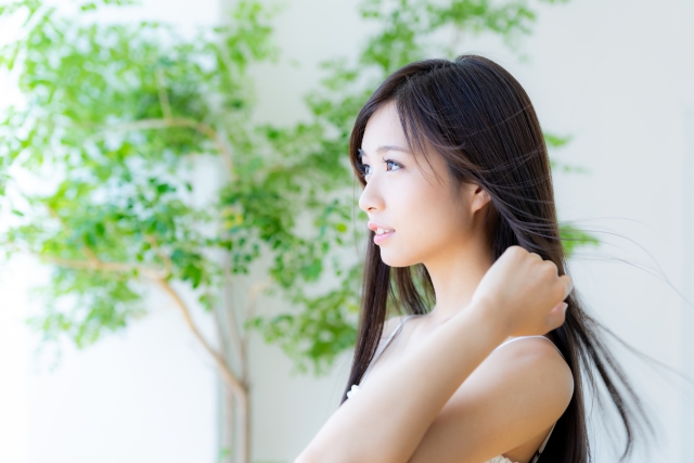 白いカーテンの窓から差し込む光の流れを受けて、顔が明るく照らされた若い女性が静かな自信を感じさせる表情をしています。背中までの長い髪の先端が軽やかな風に揺れ、髪や自己像に関する悩みを乗り越えた後の、前向きで穏やかな心境を象徴しています。