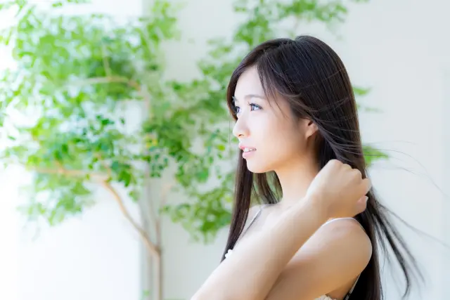 白いカーテンの窓から差し込む光の流れを受けて、顔が明るく照らされた若い女性が静かな自信を感じさせる表情をしています。背中までの長い髪の先端が軽やかな風に揺れ、髪や自己像に関する悩みを乗り越えた後の、前向きで穏やかな心境を象徴しています。