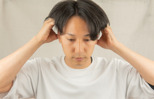 若い男性が目線を落とし、両手をこめかみに添えて髪の生え際を指先でなぞるように触れている様子。将来の薄毛や生え際の後退を気にして思い悩む様子を捉えたイメージ写真
