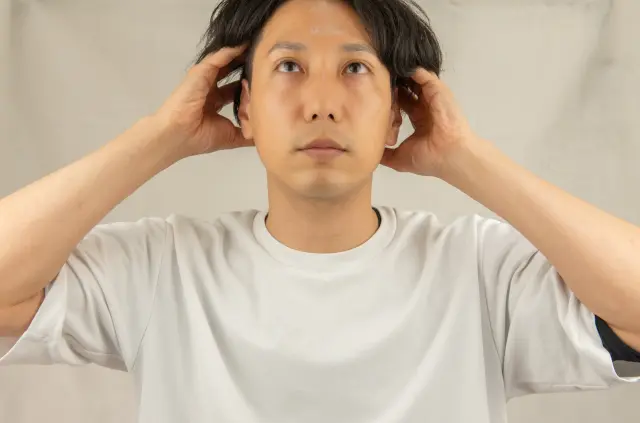 薄毛が進行し、表情も沈んだ男性が両手でこめかみ付近の髪を持ち上げている様子。頭髪のボリュームが減少し、悩みや不安を抱えている印象を与えるシーン。