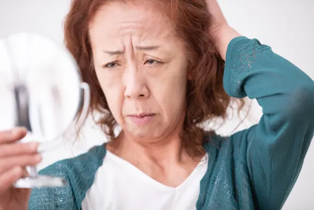 鏡を手に取り、深く眉をひそめながら自分の額を見つめる女性。後退した生え際が露出し、顔立ちは長く疲れた印象を与え、額や表情には加齢やストレスのサインがにじむ。髪のボリュームや生え際の状態が、見た目年齢や女性の自信に大きく影響することを象徴するイメージ