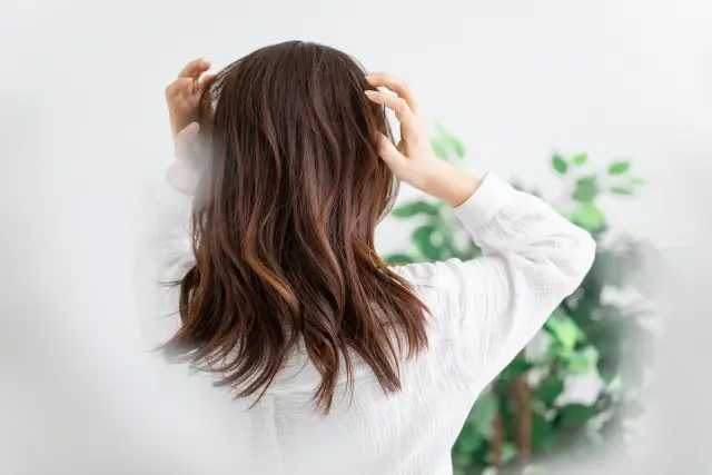 白いリネンシャツに染めた茶髪がふんわりと広がる女性の後ろ姿。室内の片隅には観葉植物が映え、リラックス感のある爽やかなシーン。髪はボリューム感があり、両手で指を通す動作が自然な美髪ケアと健康的なライフスタイルを印象づける。