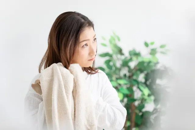 両手でタオルドライしながら長い髪を乾かす女性のイメージ。バスルームの排水口にたまる大量の抜け毛を不思議に思う様子を表現したストック写真。