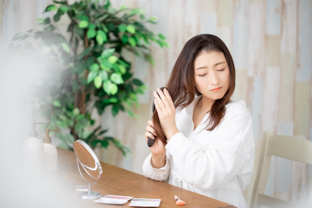 机の前で髪をとかす日本人女性。ブラッシング時の摩擦で髪が引っ張られたようで、わずかに不快そうな表情を浮かべている。摩擦ダメージや絡まりによるヘアトラブル、日常のヘアケアの注意点を示すシーン。