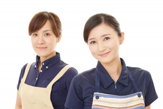 エプロンを着けて並んで微笑む40代後半の女性2人のイメージ写真。家庭的で温かみのある雰囲気が伝わるシーン。年齢を重ねた女性の健康や美容意識、自然体の美しさを象徴する写真として使用可能。