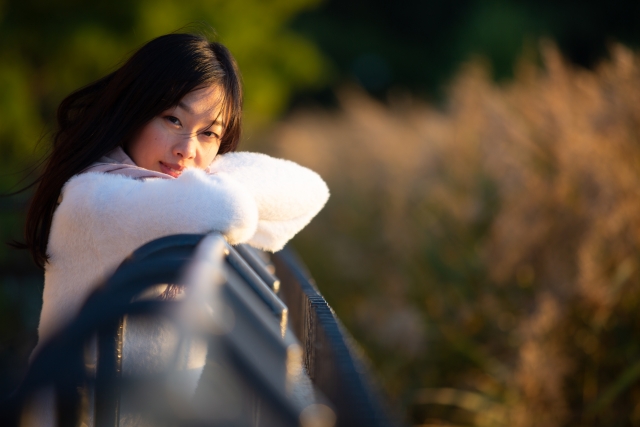 公園の手すりに腕と上半身を預けて佇む女性。日差しが黒髪の頭頂部に当たり、艶やかに反射している。