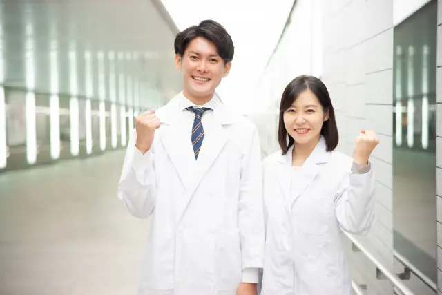白衣をまとった男女の医療従事者が、力強く拳を握り希望と決意を込めて微笑む姿。明るさの中に確かな専門性と信頼感が漂い、「知識は金にまさり、技術は精錬された銀にも勝る」という信念を体現する一枚。誠実な医療の力強さと安心感を伝えます。