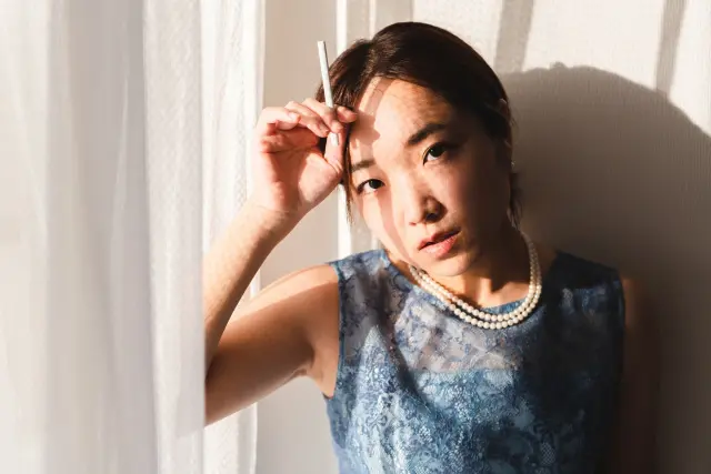 窓際に座り、昼のカーテン越しの光を背にたたずむ女性|青い大理石模様のノースリーブドレスをまとい、上を見上げる表情は無言の感情を湛え、眉に手を添えた姿が印象的。指には点火されていないタバコが挟まれ、空虚と静寂を象徴するような一瞬を切り取ったイメージ