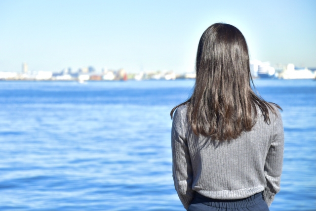 港の桟橋で穏やかな海に向かう女性の後ろ姿 — 柔らかな陽射しを受けて輝く黒髪が、青空と静かな海を背景に美しく際立ち、東アジア系の髪質と染毛の特性を象徴する情景