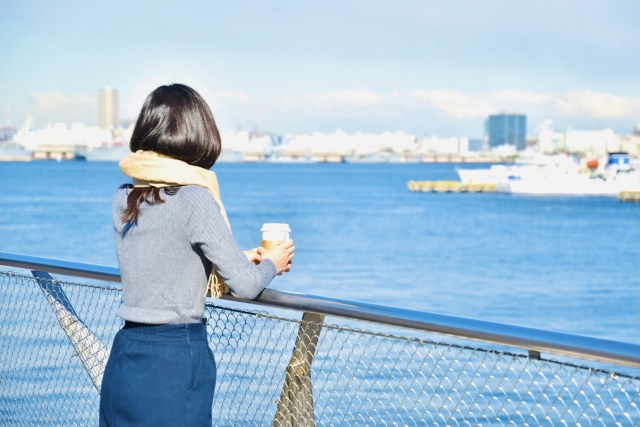 港で青い海を眺めながら紙カップを手にする黒髪の女性。髪が太陽の光で艶やかに輝き、健康的な髪の印象が自己イメージや自信にもつながることを想起させるリラックスしたワンシーン。