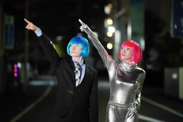 近未来的な衣装に身を包んだ男女が、人気のない夜の街角で街灯の光に照らされながら空を指さしている姿。カラフルなウィッグをかぶっているが、その表情には「いつか本物の髪が頭皮から生える日が来る」という希望が込められている。人類が月へ到達したように、薄毛や脱毛の悩みも医療技術の進歩によって克服できる未来を象徴するビジュアル。毛髪再生医療、自毛植毛、先端発毛技術の未来を連想させるイメージ。