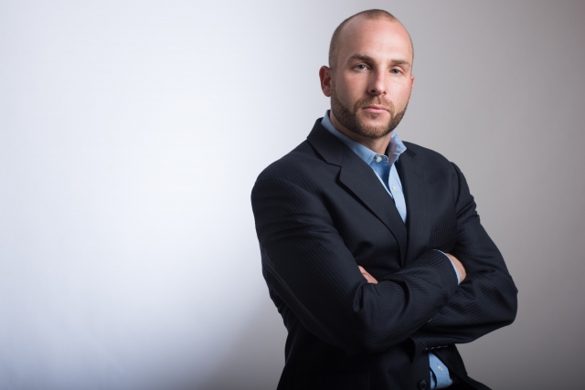 スーツ姿で腕を組み、自信に満ちた表情を浮かべる中年男性。額の生え際が後退し、頭頂部の髪が薄くなってきているが、それを堂々と受け入れている様子。口元やあご周りには濃いヒゲがあり、加齢による男性型脱毛症（AGA）の進行を自分らしく受け止めた姿。薄毛でも自信を持ち、生き方を変えない現代男性のイメージ。