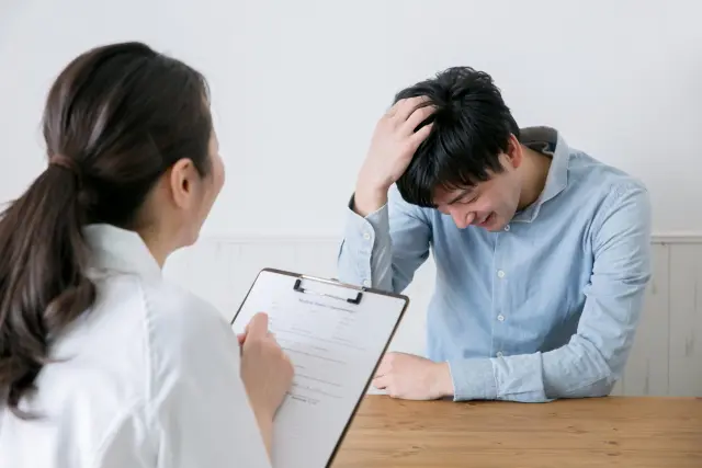 植毛を検討中の男性が、薄毛の状態を看護師に相談しながら頭頂部に手を添えている様子。FUE植毛のカウンセリング場面をイメージしたシーン。