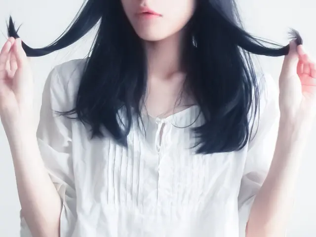 白いリネンシャツを着た女性が黒髪に手を添え、わずかに開いた唇と明るい肌が髪の艶とコントラストを描き出す。髪の質感・色彩・静電気による広がりの対比が美しさの一部として視覚化された場面。