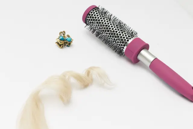 ブロンドのカールがかかったヘアエクステンションと、ピンクの獣毛タイプのブラシ、小さな金属製のヘアクリップが並ぶ様子。非外科的増毛で使われる人工毛やケア用アイテムをイメージしたスタイリングツールの一例。