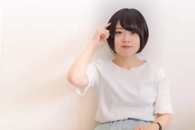 こめかみの髪を指さしながら頬をふくらませるボブヘアの小柄な女性。自毛植毛後の生え際や側頭部の髪の量に悩みつつ、スタイリングや分け目の工夫を検討している様子を表現