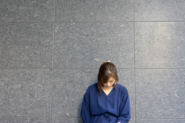 灰色の大理石の壁にもたれ、うつむく女性の後ろ姿。距離があっても頭頂部の地肌がはっきりと見えるほど、薄毛が進行している様子が印象的。周囲に気づかれやすい女性の脱毛の悩みを象徴する場面。