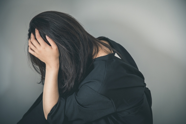 黒いドレスを着た女性が顔を手で隠すように体を傾けて座る姿。事故や外傷による目立つ瘢痕や脱毛は、外見だけでなく心理的にも深い影響を与えることを象徴。無償植毛支援「オペレーション・リストア」により自信を取り戻す希望の文脈を表現。