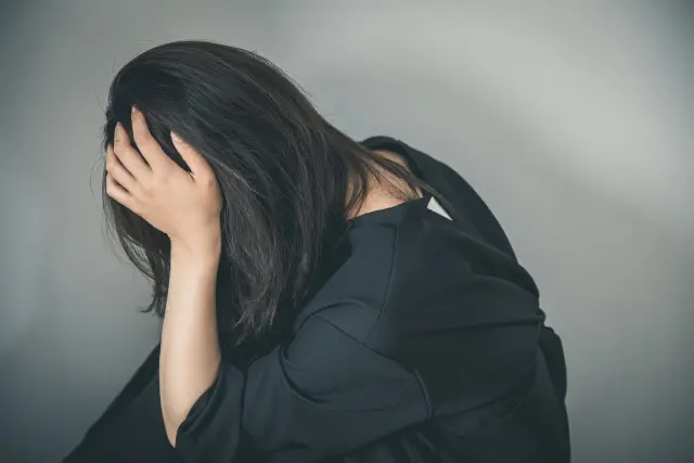 黒いドレスを着た女性が顔を手で隠すように体を傾けて座る姿。事故や外傷による目立つ瘢痕や脱毛は、外見だけでなく心理的にも深い影響を与えることを象徴。無償植毛支援「オペレーション・リストア」により自信を取り戻す希望の文脈を表現。