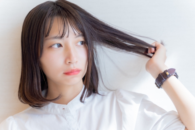 肩まで伸びた黒髪を無意識に触れる女性の横顔。艶やかな髪が顔の輪郭を際立たせ、整った眉毛と上品にカールしたまつげがアーモンド形の目元を美しく引き立てている。髪や眉毛、まつげといった顔の毛が、外見の印象や表情の柔らかさに大きな影響を与えることを象徴するイメージ。