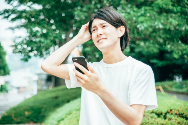 風が吹く公園を歩く若年男性。頭頂部の地肌が露出しはじめた部分に手をやりながら、スマートフォンで薄毛の情報を検索する姿。脱毛の進行に不安を感じ、戸惑いの表情で空を見上げる瞬間。