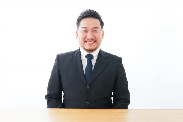 スーツ姿の中年男性がデスクに座り、穏やかな笑顔で前向きな印象を与える様子。自然な生え際や毛髪の印象がビジネスシーンでの自信や清潔感に影響することを表現したイメージ。