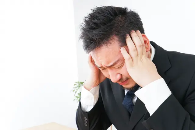 中年の日本人男性が頭を両手で抱え、薄毛による見た目の変化と社会的なプレッシャーに苦悩している様子。髪の悩みに対する職場での偏見や通勤時の周囲の視線、女性同僚の陰口に深く傷つき、自尊心の低下とストレスを抱えるミドル世代男性のリアルな心情を象徴するイメージ。薄毛・脱毛による心理的影響や社会的差別を扱うコラム記事の文脈に沿った、精神的負担と孤独感を表現したビジュアル。