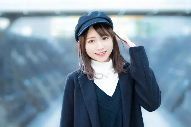 紺色のキャップとセーターを身に着けた若い女性が、帽子のつばに軽く手を添えて挨拶するような仕草を見せる姿。髪を帽子でカバーするスタイルは、女性の薄毛をさりげなく隠す選択肢としても用いられることがある。