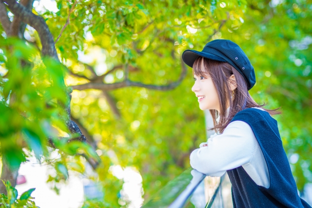 木漏れ日の差し込む森の公園で、青い帽子をかぶった若い日本人女性が、手すりにもたれて穏やかな笑顔を見せる様子。抗がん剤治療中でも自然の中でリラックスし、自分らしさや心の安らぎを大切にする姿をイメージさせるシーン。
