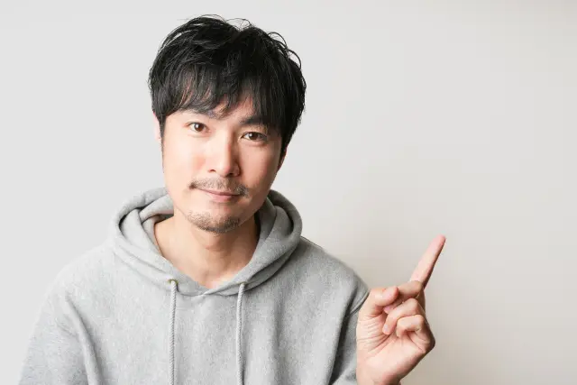 リラックスした姿勢で椅子に座りながら上を指さす男性。穏やかな表情の中に前向きな意志を感じさせ、若年性薄毛(AGA)に悩みながらもポジティブな視点で将来の選択肢を模索する男性像を象徴している。