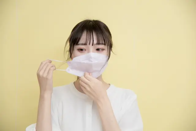 マスクをゆっくりと外しながら、不安げな表情を浮かべる若い女性。薄毛や抜け毛の悩みが外見への自信や心の不調に影響を与える様子を表現したイメージ。
