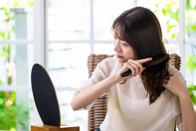 小さな鏡の前でデスクに座り、両手で丁寧に髪を整える女性。健康的な髪を保つには、毎日のヘアケア習慣が重要であることを示す日常の一コマ。