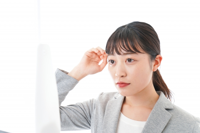 無表情のまま前髪を整える女性が吊り鏡を見つめる場面。まつ毛に特別な特徴はなく、目元の印象が控えめなことが、まつ毛や眉毛が顔全体の印象に与える役割や、自然な再建手術の必要性を静かに示唆しているビジュアル。