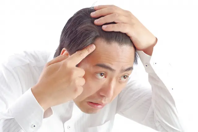 男性が左手のひらを頭頂部にあて、右手で生え際をつつきながら、真剣なまなざしで鏡を見つめている。表情はまるで魚のように緊張感が漂う。