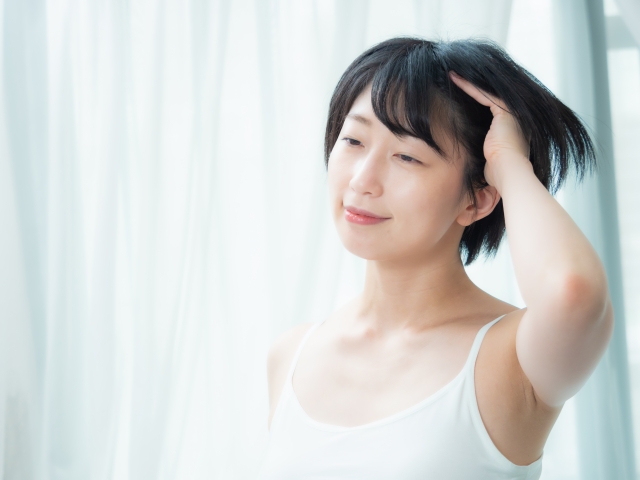 白いキャミソールを着た女性が黒髪に指を通す様子。毛髪の成長や抜け毛が気になる読者に向けた、毛周期やヘアケアの重要性を示すイメージカット。