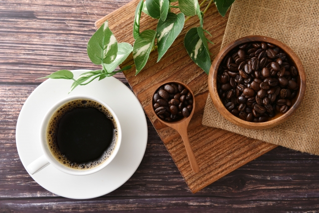 白いカップとソーサーに注がれたブラックコーヒーとコーヒー豆、フェイクグリーンや木製素材で演出されたウッディなテーブル上の美的配置｜自然素材とコーヒーの美容・育毛効果を特集するヒロクリニック監修コラム用イメージ