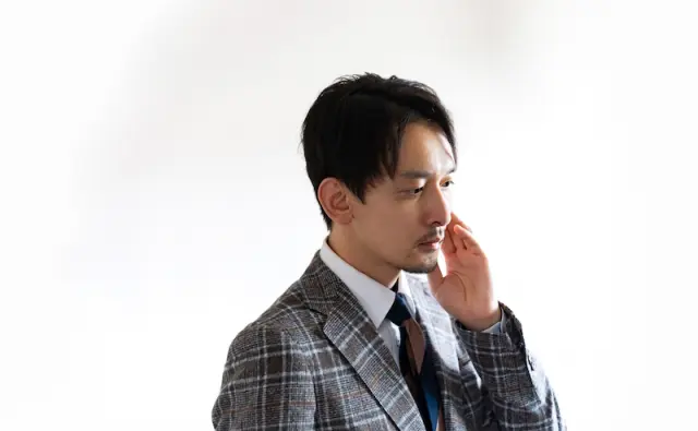 左手でヒゲの生えた頬に軽く触れる日本人ビジネスマン。口ひげとあごひげが丁寧に整えられており、清潔感と男らしさを兼ね備えた都会的な印象を与える。都市部では珍しいヒゲスタイルを持つ男性の例として、ヒゲ移植による外見変化や自己表現の可能性を象徴するイメージ。