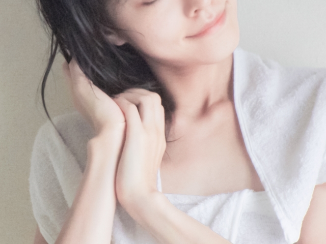 お風呂上がりの女性がウールタオルで黒髪を乾かしているシーン。微笑みを浮かべた口元が印象的。健康な髪は多くの人が当たり前に思っているが、失って初めてその大切さを実感することが多い。