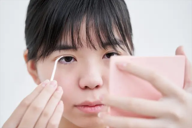ピンクの手鏡を見ながらまつ毛を整える日本人女性。まつ毛の美しさや日常的なケアの重要性を示すワンシーンで、美容目的のまつ毛移植の関心にもつながるイメージ。