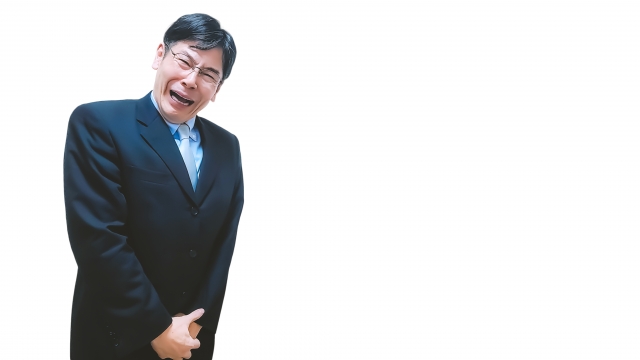 中年のビジネスマンが絶望的な表情で股間を両手で押さえ、口を開けてうめく様子。フィナステリド服用後の思わぬ副作用である性機能障害に衝撃を受けた男性のイメージ。AGA治療とPFS（ポスト・フィナステリド症候群）のリスクに関連した場面を象徴