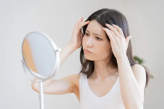 前髪に違和感を覚えながら鏡の前で髪を確認する女性。薄毛やボリューム不足に気づき始めた初期段階の不安を象徴する場面｜女性の薄毛や自毛植毛に関する情報コラム用画像