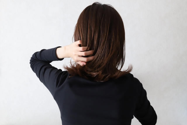 髪の後頭部に左手を添えるように肘を上げた女性の後ろ姿。頭皮や髪の状態を自ら確認する仕草は、髪のパサつきや抜け毛、牽引性脱毛症といったトラブルへの不安や関心を表しており、記事内で紹介される「髪の変化が知らせる体の不調サイン」というテーマと視覚的にリンクする構成。