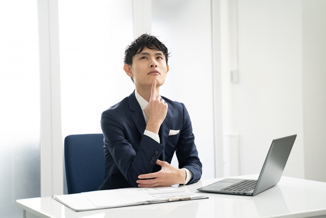 ノートパソコンの前で顎に指を添え、真剣な表情で悩む若年の日本人ビジネスマン。将来や外見への不安、若年性脱毛症の心理的影響を象徴するイメージ。