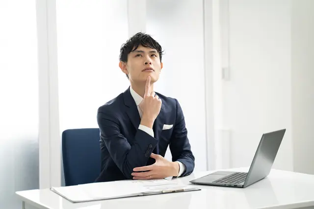 ノートパソコンの前で顎に指を添え、真剣な表情で悩む若年の日本人ビジネスマン。将来や外見への不安、若年性脱毛症の心理的影響を象徴するイメージ。