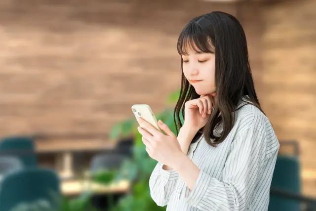 髪の健康に関するコラム記事をスマートフォンで真剣に読みながら、右手の拳に顎を乗せて思案する若い女性。抜け毛や白髪など髪の変化と体調の関係に関心を寄せている様子がうかがえる構図で、記事のテーマである「髪のSOSサイン」やホルモンバランスの乱れに関する話題と関連性が高い読者の共感を誘うイメージ。