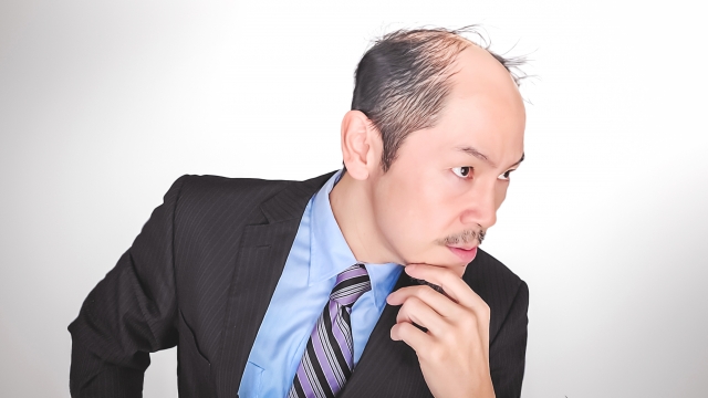 頭頂部から前頭部中央にかけて広範囲に脱毛が進行し、左右の髪が孤立して見える若年男性。無表情で前方を見つめ、年齢以上に老けた印象を与える。若年性脱毛症の進行例と心理的影響を象徴するイメージ。