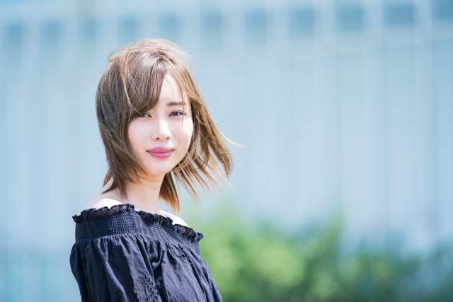 屋外で微笑みながら立つ女性。明るい茶色に染められた髪が、通り過ぎる風にそよぎ、自然な美しさを引き立てている。風になびく髪は、その人の柔らかい印象や健康的な雰囲気を演出し、髪型が見た目や自信に与える影響の大きさを感じさせる一瞬。