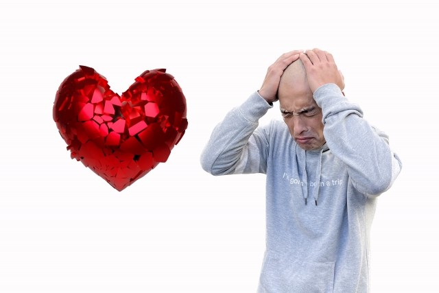 脱毛に悩む男性が頭を抱え、悲しげな表情を浮かべている。隣にはひび割れたハートのイラストが描かれ、若年性脱毛症による自己肯定感の低下や恋愛関係の喪失を象徴するイメージ。