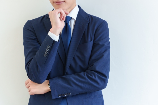 ビジネススーツ姿の男性が腕に顎を乗せて思索的な表情を見せる構図。顔の上半分が隠れており、生え際やヘアラインの印象が隠されていることで、外見の一部が見えないことが自己認識や自信に影響するという文脈を示唆するイメージ。